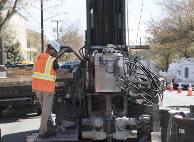 Pictured: A driller in a hi-viz vest and hard hat stands at the controls of a sonic drilling rig on a city street.
